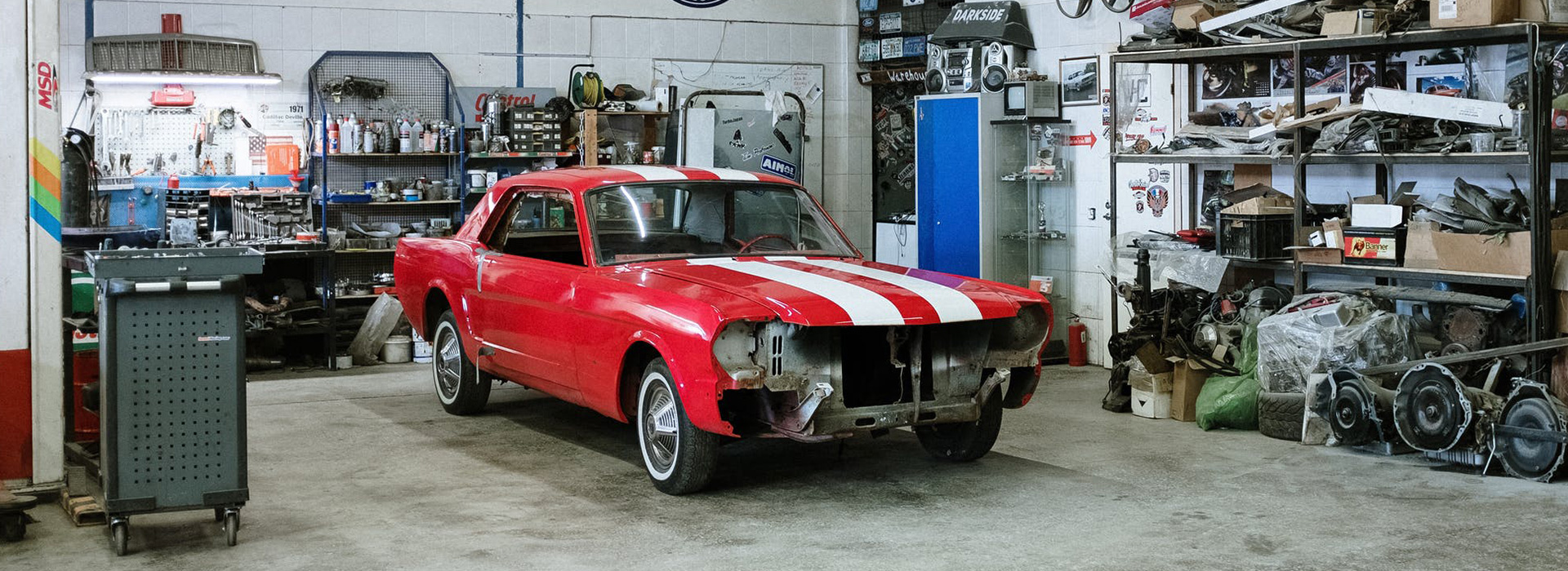 L'image représente une voiture rouge ornée de bandes blanches. Elle est garée à l'intérieur d'un atelier de réparation automobile, entourée de diverses pièces et équipements automobiles.