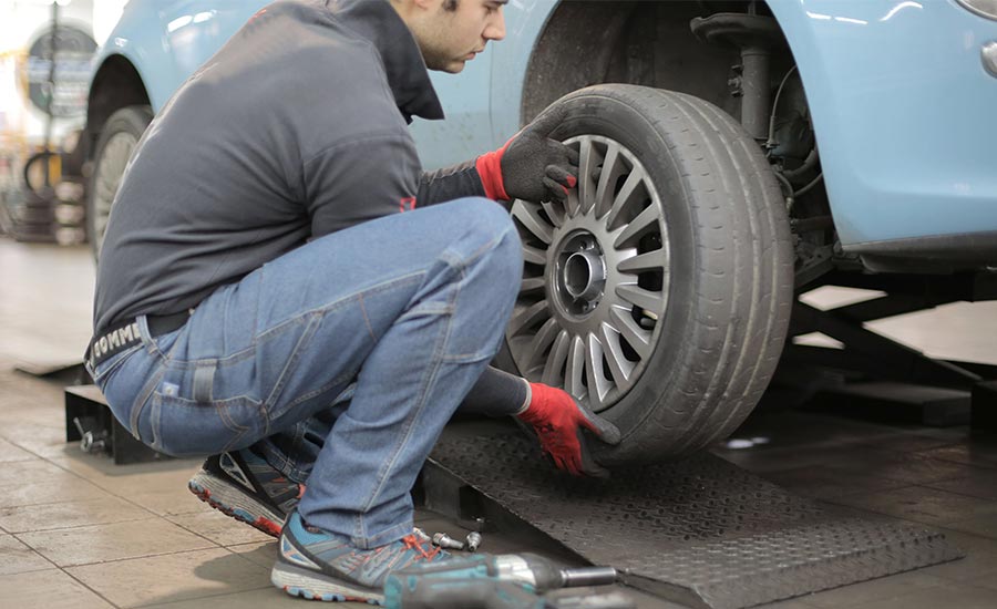 Un homme travaille sur un pneu dans un atelier de réparation automobile. Il porte un jean et des chaussures adaptées à sa tâche. L'environnement semble être en plein air, avec un accent sur le système de roue automobile et ses composants.