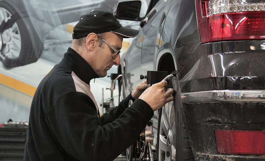 Un homme travaille sur une voiture, se concentrant sur les pneus et les pièces automobiles. Il porte des vêtements adaptés au travail mécanique et se trouve dans un atelier en plein air. Des outils se trouvent à proximité tandis qu'il s'occupe du véhicule.