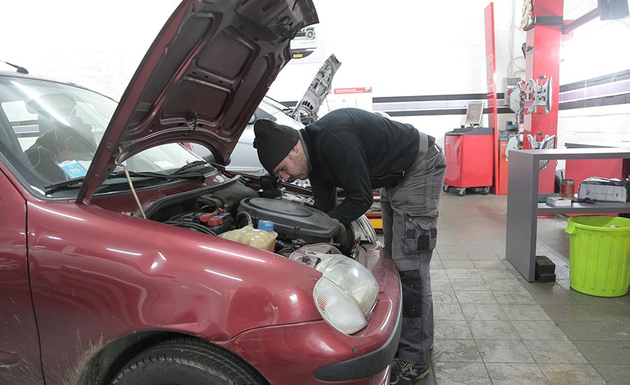 Un homme se tient à l'extérieur et regarde le capot d'une voiture rouge garée. La scène suggère qu'il est en train d'inspecter ou de diagnostiquer un problème avec le véhicule. Cette image présente des éléments liés à la réparation automobile, notamment des pneus et des roues associés à des véhicules terrestres.