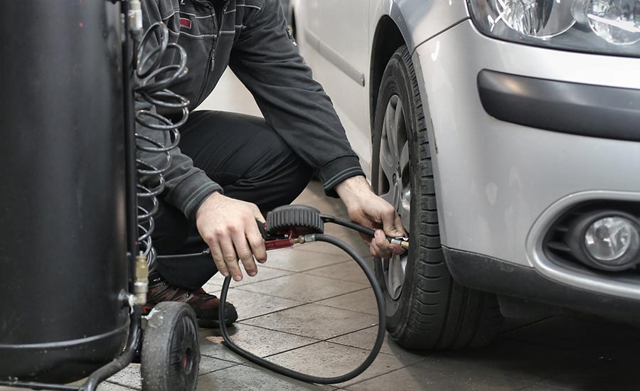 Un homme utilise une pompe à air pour gonfler un pneu. Il est à l'extérieur, debout sur le sol, et porte des vêtements et des chaussures décontractés. La scène présente des éléments liés à un véhicule, notamment une roue et un pneu d'automobile.