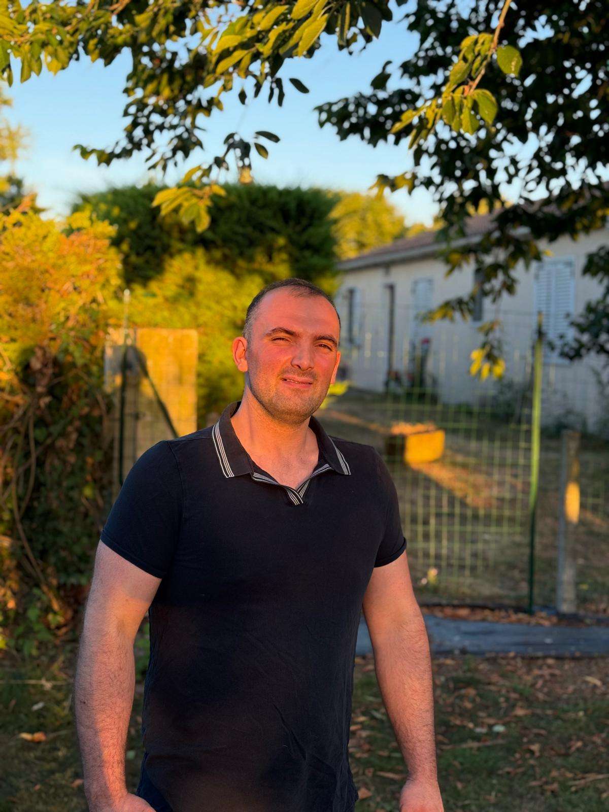 Un homme se tient à l'extérieur, au soleil, et sourit. Il est entouré d'arbres et d'un ciel dégagé. La photo capture son attitude joyeuse tout en mettant en valeur ses vêtements et ses traits.