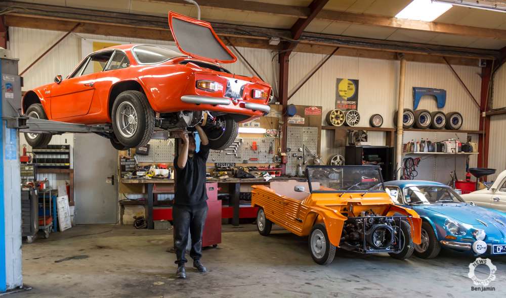 Un homme est représenté tenant une voiture à l'intérieur d'un atelier de réparation automobile. Le décor comprend diverses pièces et composants automobiles, le véhicule reposant sur le sol. L'environnement semble être un espace industriel intérieur, caractérisé par ses couleurs orange et sa structure de plafond visible.
